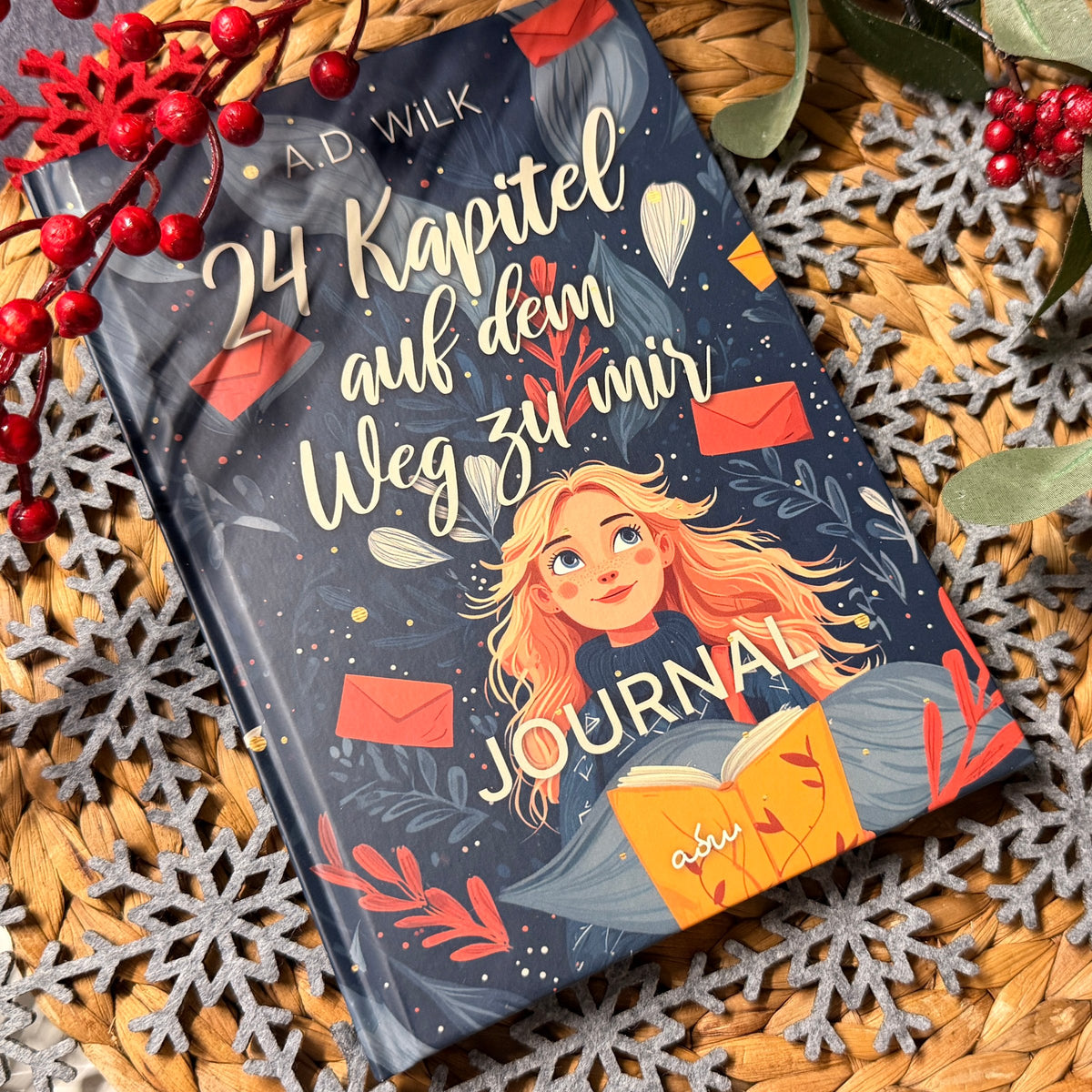 Journal zu »24 Kapitel auf dem Weg zu mir« von A.D. WiLK, Hardcover, auf einem Rattan-Untersetzer mit roten Beeren und Filz-Schneeflocken.
